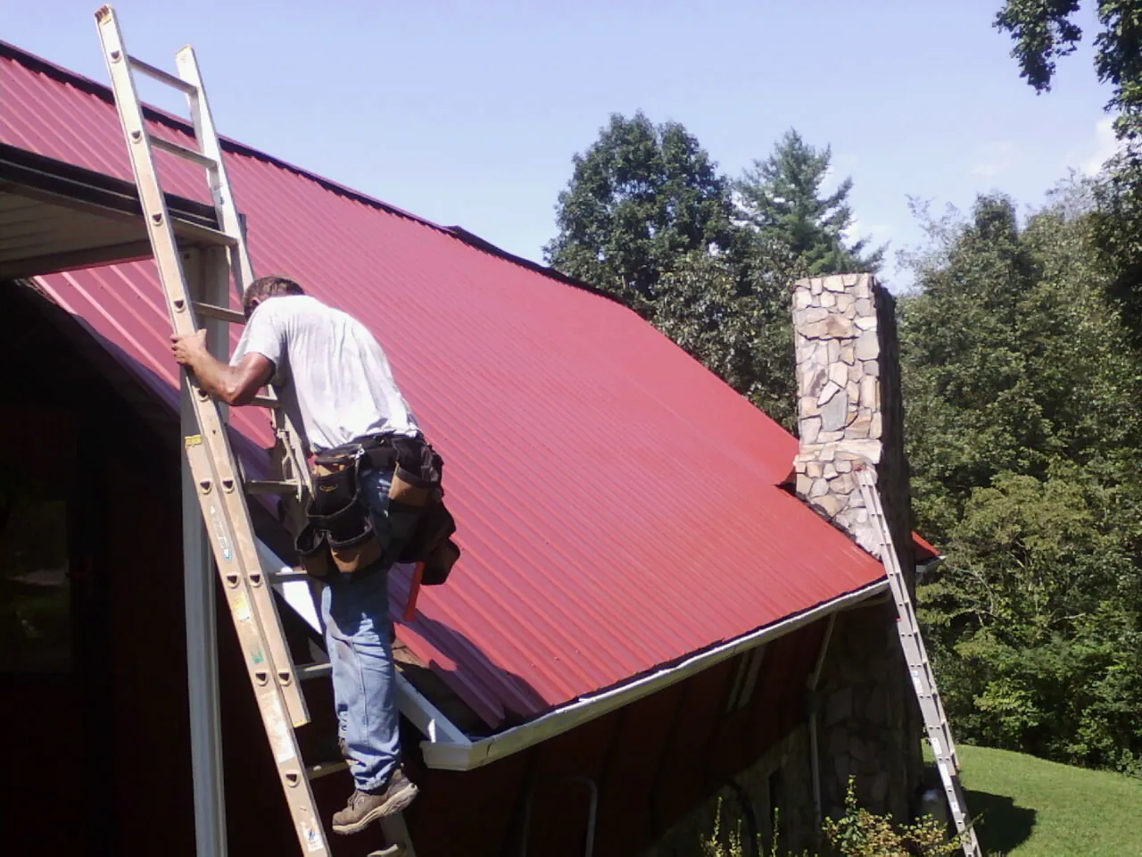 Roofer on a ladder beside a red metal roof and stone chimney — Soffit & Fascia Repair by Roofing Brothers in Shreve